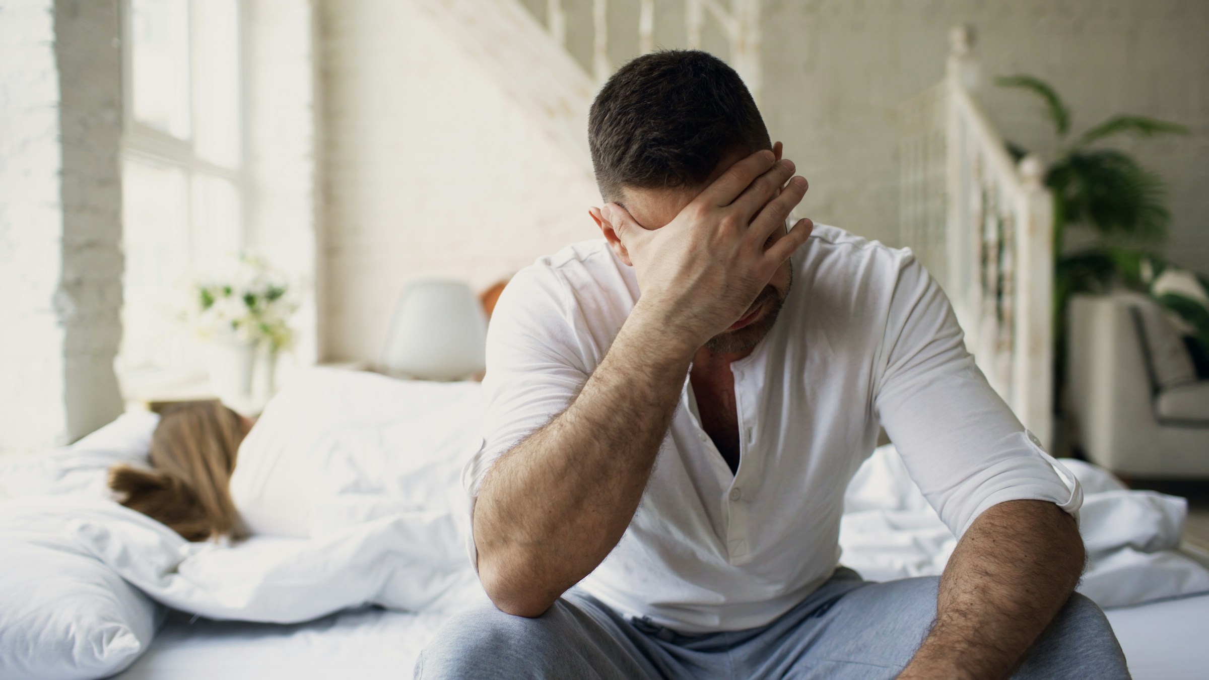 A man sitting on the edge of a bed with his hand pressed to his face