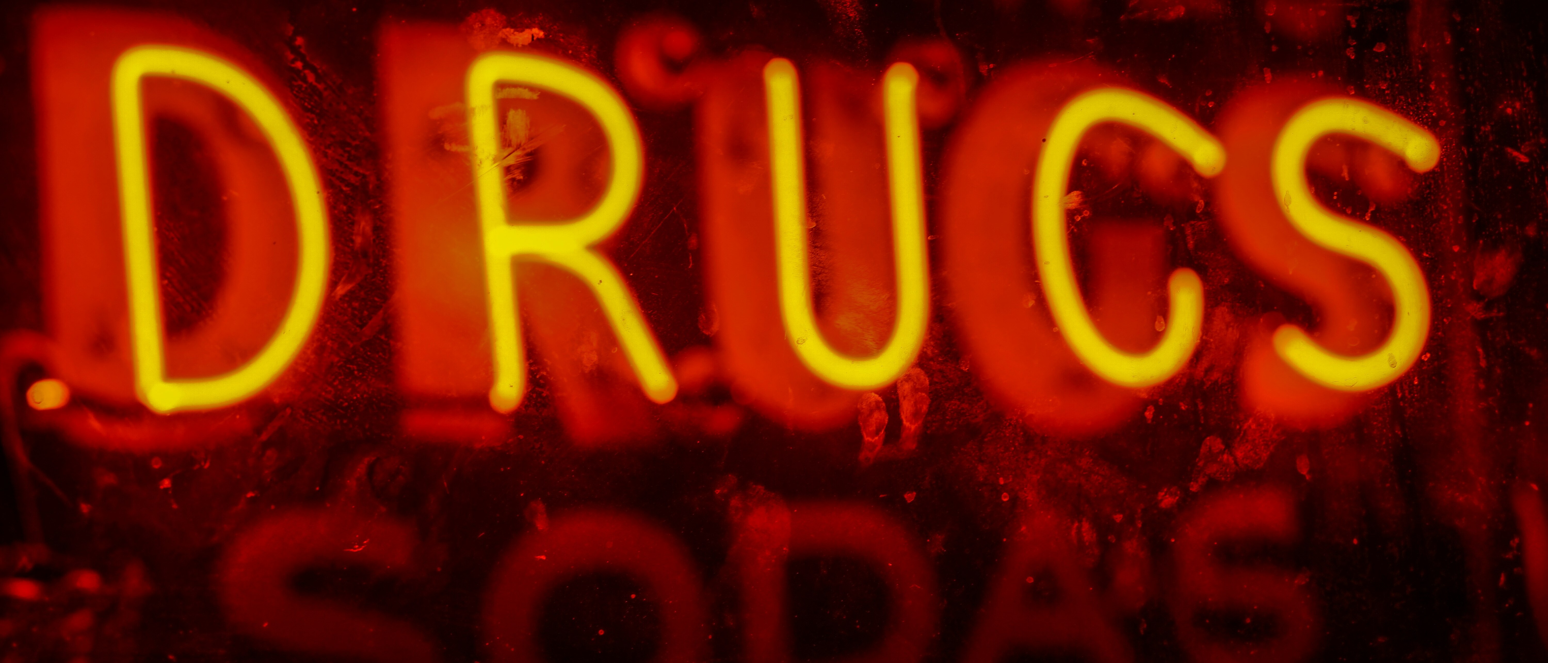 A glowing yellow neon sign reading DRUGS against a deep red, weathered backdrop