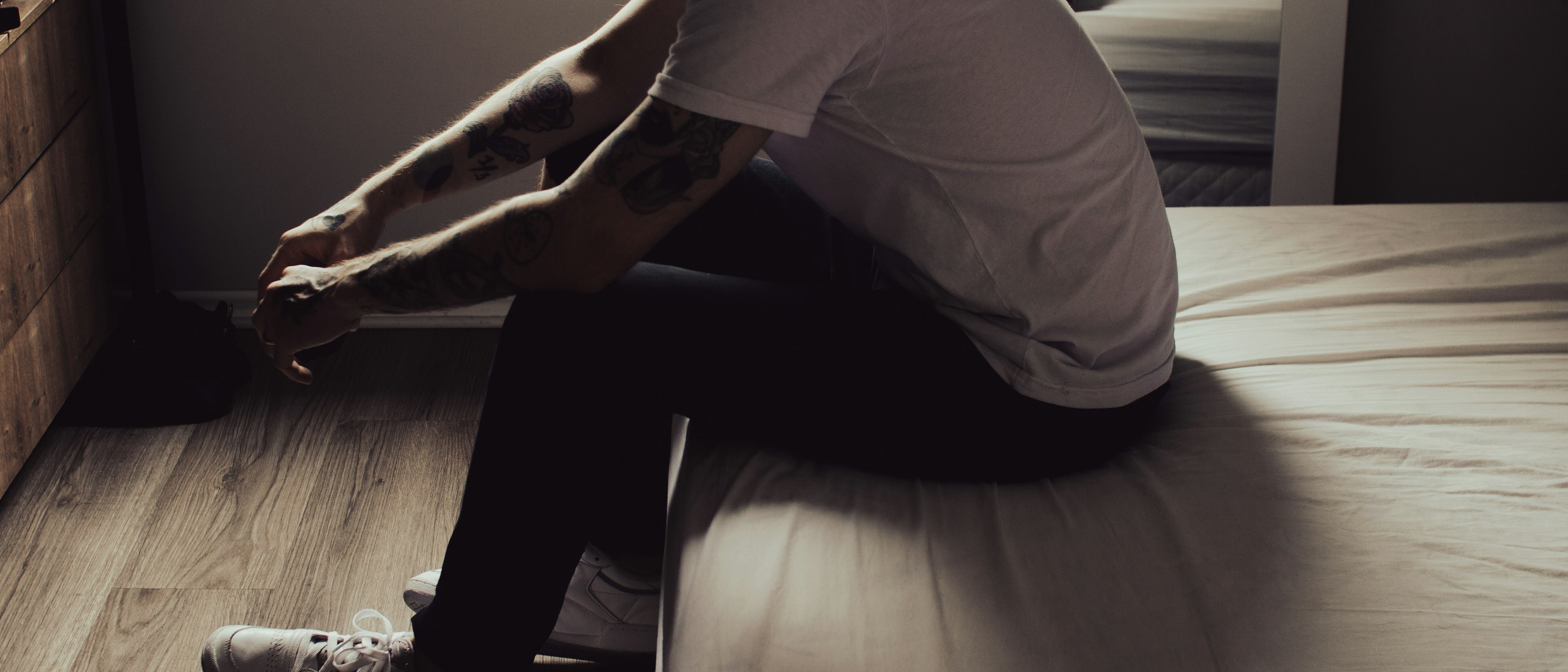 A man sitting on the edge of a bed in soft side light, head out of frame, hands clasped between his knees