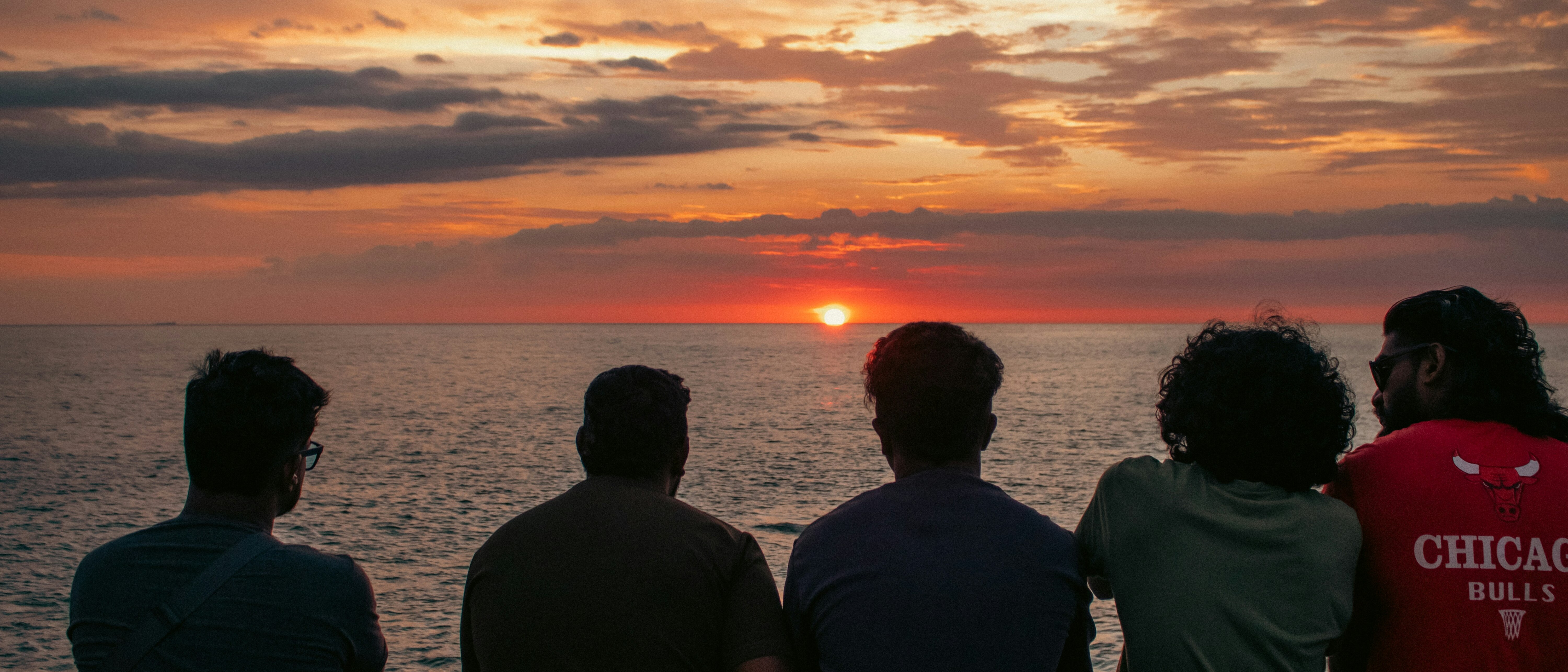 Five men sitting in a line watching a sunset over the sea, from behind