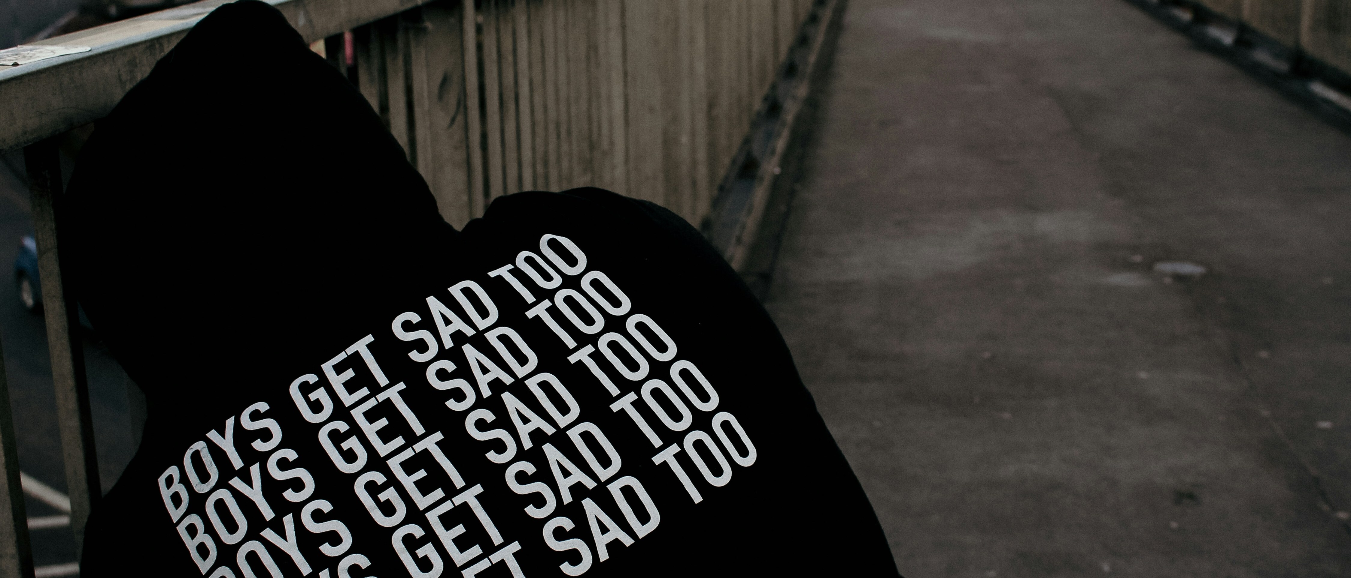 A hooded figure on a footbridge wearing a sweatshirt reading BOYS GET SAD TOO